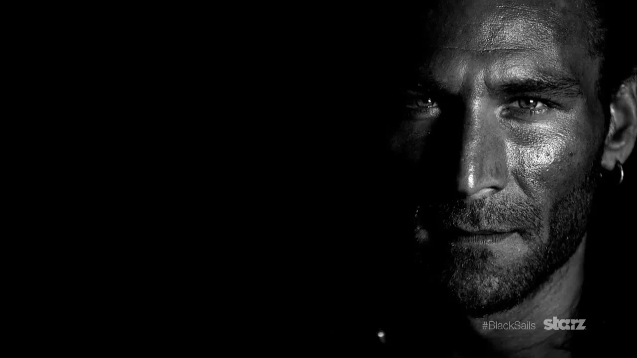 A man with a beard and an earring is shown in a dramatic closeup, with half his face in shadow, against a black background.