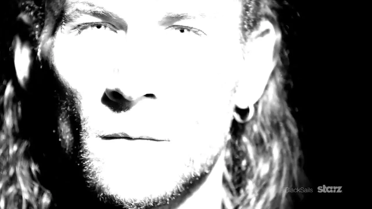 A man with long hair and a beard looks intensely at the camera in a black and white, high-contrast shot.