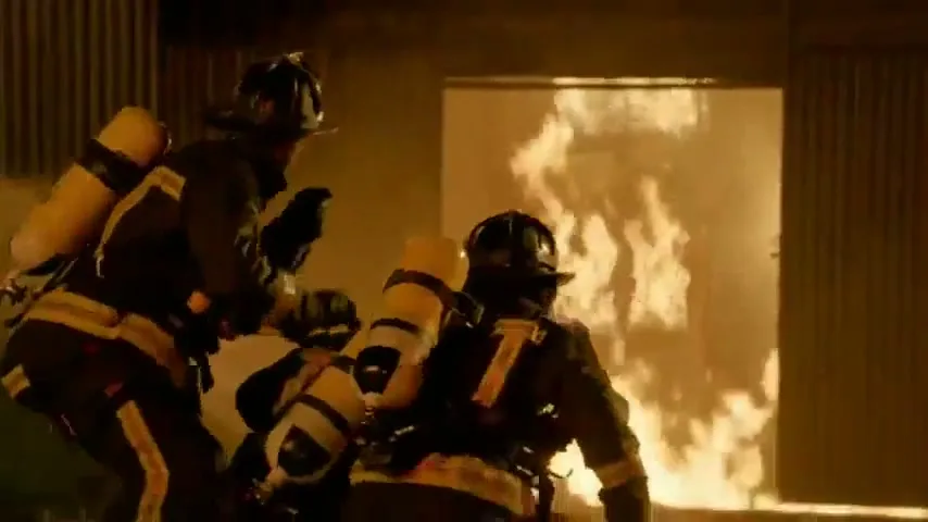 Two male firefighters in full gear and oxygen tanks enter a burning building, with flames visible in the doorway.