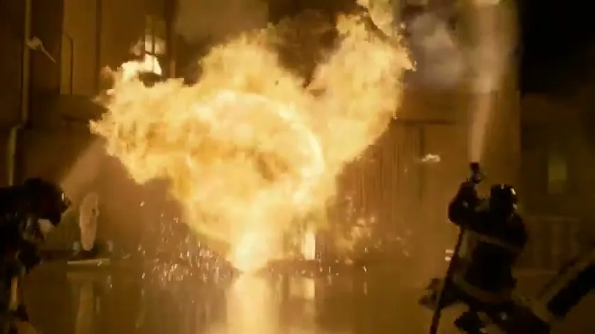Firefighters in dark gear battle a massive explosion of fire erupting from a building at night, with debris flying.