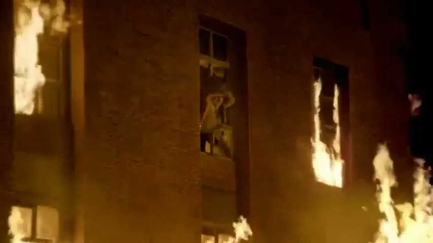 A woman in a white dress and a man in a red shirt are trapped in a burning building, looking out a window in distress.