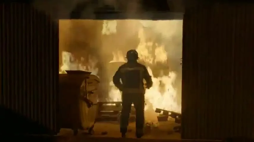 A male firefighter in full gear stands in a doorway, looking into a room engulfed in flames and smoke.