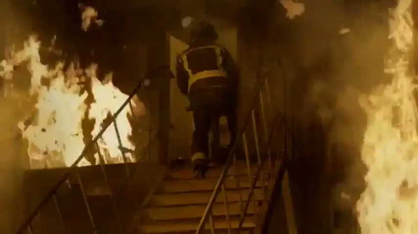 A male firefighter in full gear walks up a burning staircase in a building engulfed in flames.