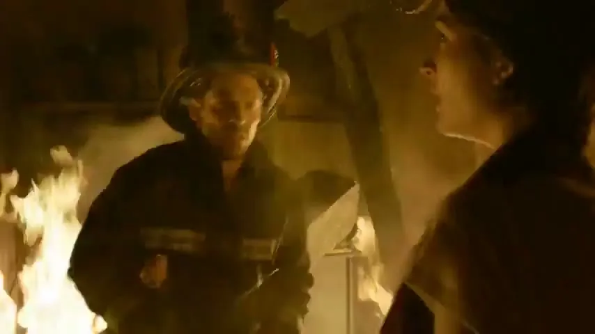 A male firefighter in a burning building removes his helmet and jacket, revealing a white t-shirt, as a woman looks at him intently.