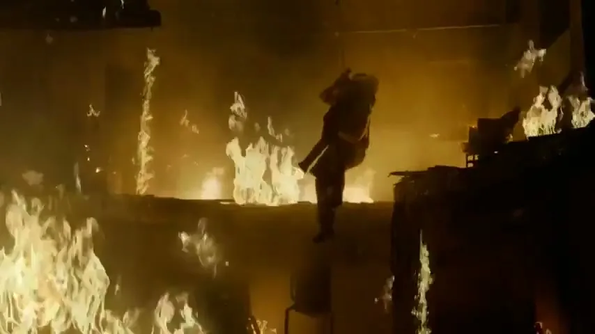A fireman in full gear carries a person through a burning building, with flames engulfing the structure around them.