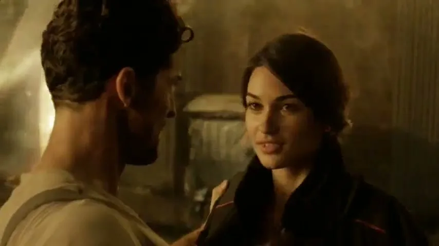 A man and woman stand close, looking at each other, as the woman speaks to the man in a smoky, industrial setting.