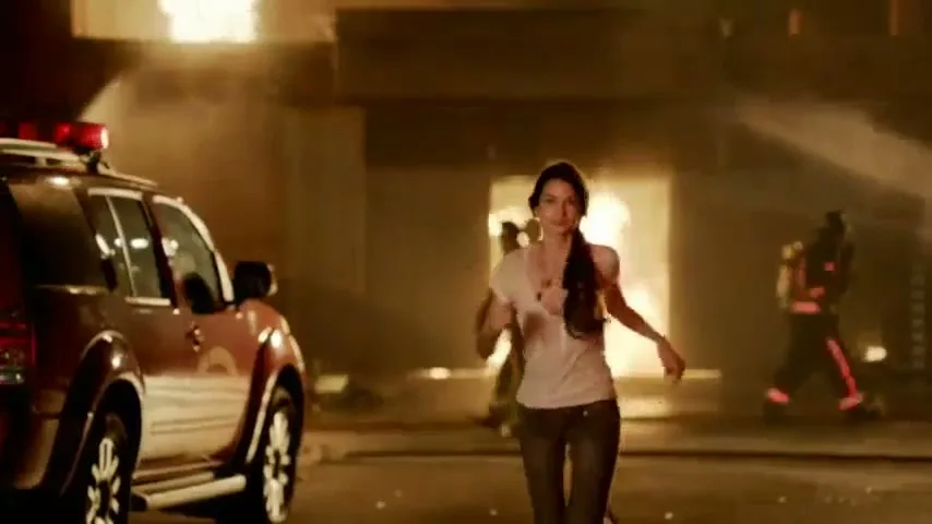 A beautiful woman in a white shirt and dark pants runs towards the camera, with a fire truck and firefighters in the background of a burning building.