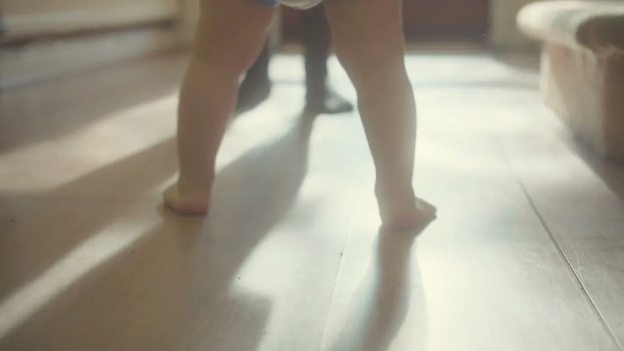A baby's bare legs and feet are seen from behind as they stand on a light wooden floor, with an adult's feet visible in the background.