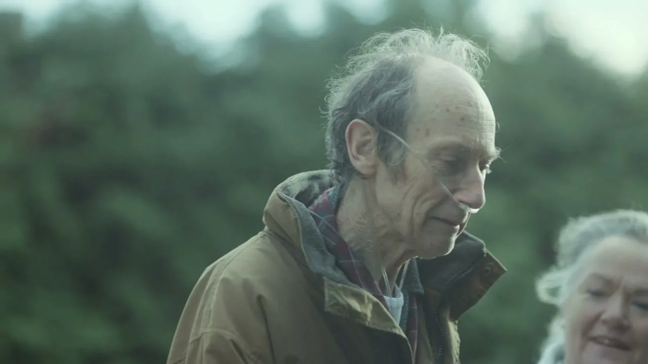 An elderly man with an oxygen tube in his nose, wearing a brown jacket, looks down and smiles slightly, with an elderly woman out of focus behind him.