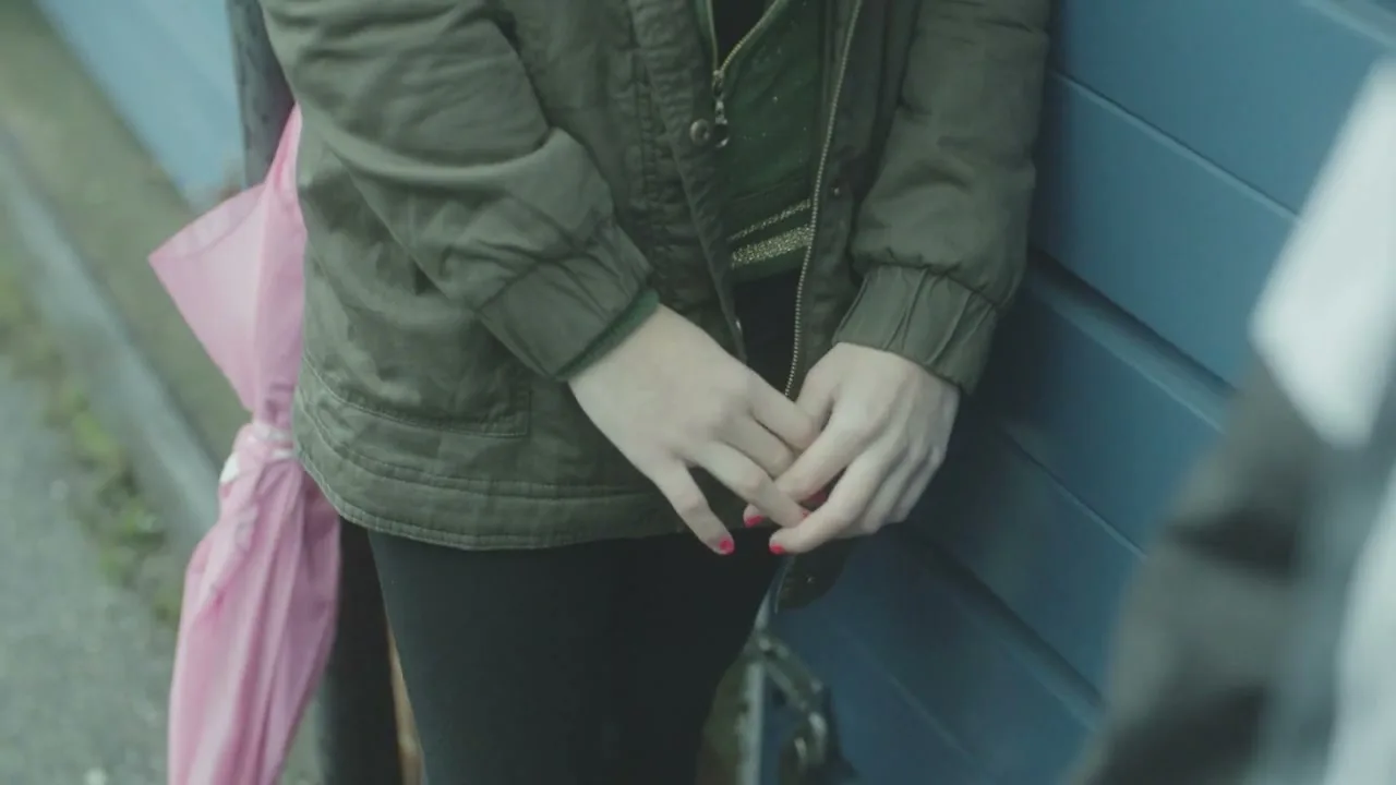 A young person in a green jacket and black pants stands with their hands clasped, leaning against a blue wall with a pink umbrella beside them.