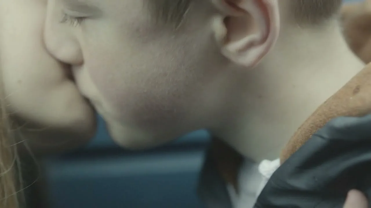 A young boy with short hair and a woman with long hair share a tender kiss on the cheek, in a close-up shot.