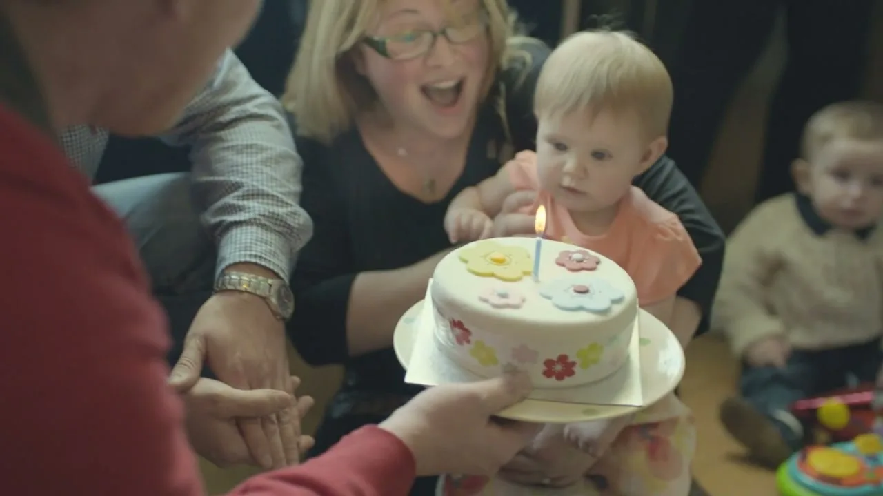 A group of adults and babies are gathered around a small birthday cake with a single candle, celebrating a baby's first birthday in a cozy room.