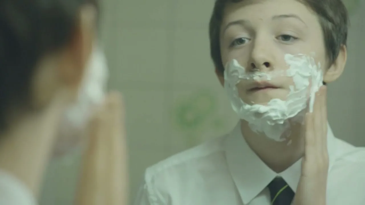 A young boy with short brown hair, wearing a white shirt and striped tie, applies shaving cream to his face while looking into a mirror in a bathroom.