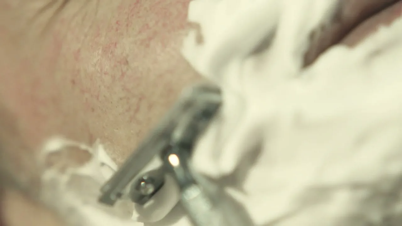A close-up shot shows a person's neck being shaved with a safety razor, removing white shaving cream.
