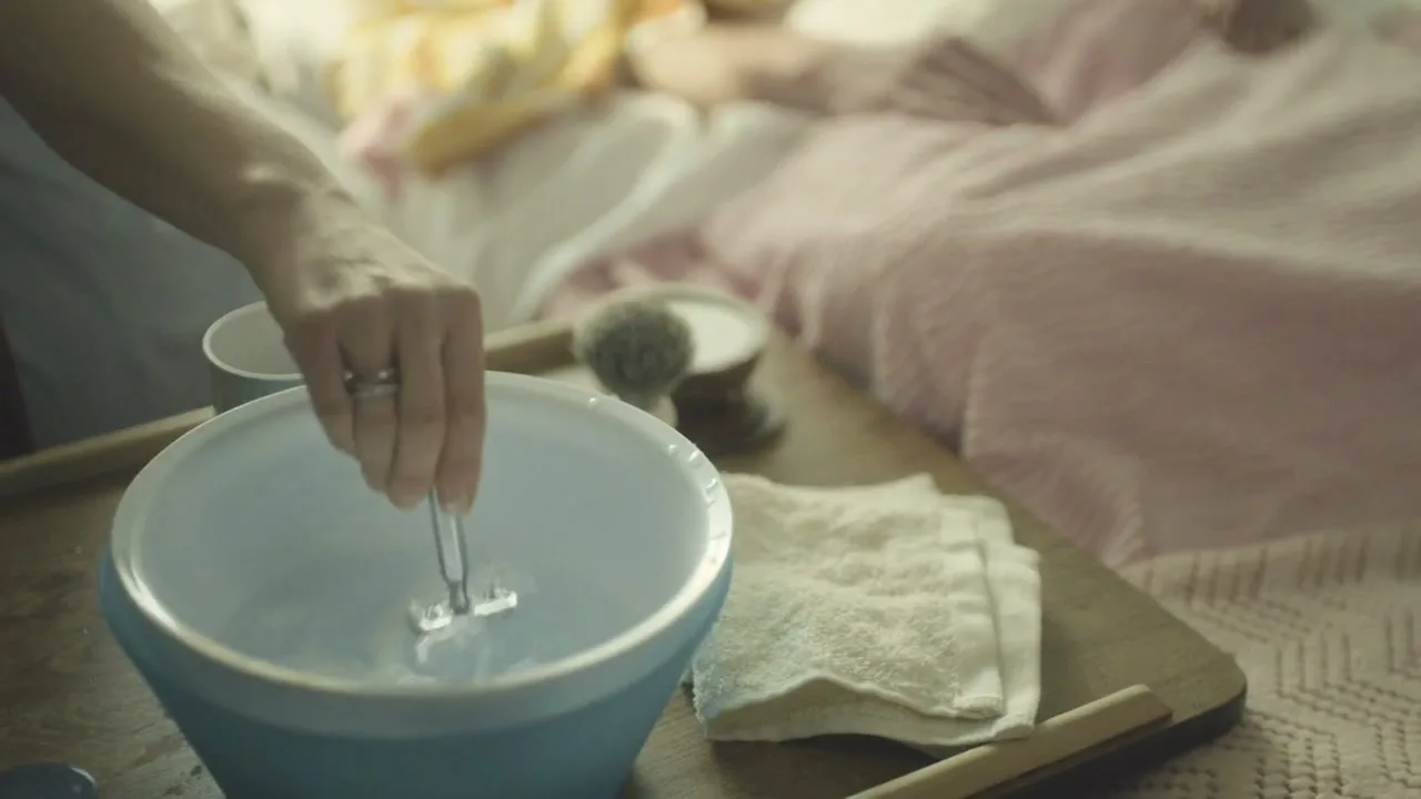 A person's hand dips a razor into a blue bowl of water on a wooden tray next to a bed, where another person is lying down.