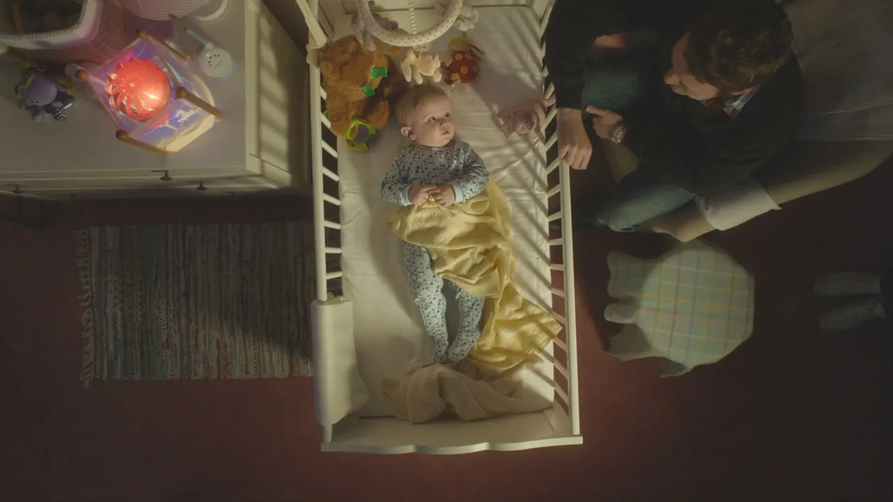 An overhead shot shows a baby lying in a crib, looking up, while an adult sits beside the crib, looking down at the baby.