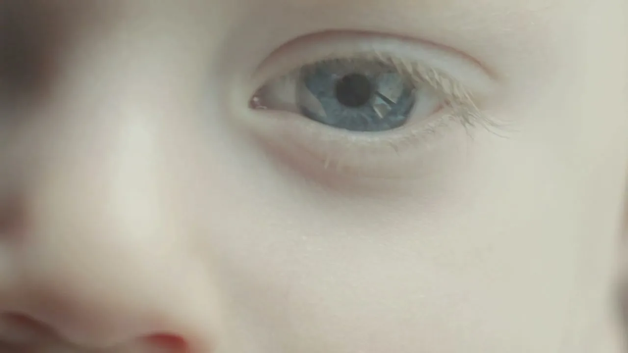 A child's face is shown in a close-up, with their blue eyes looking directly at the camera, in an interior setting.