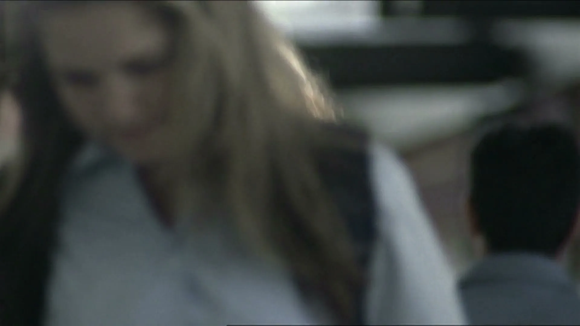 A diverse group of people walk through a busy, modern public space, with a woman in a grey shirt and dark hair looking directly at the camera.