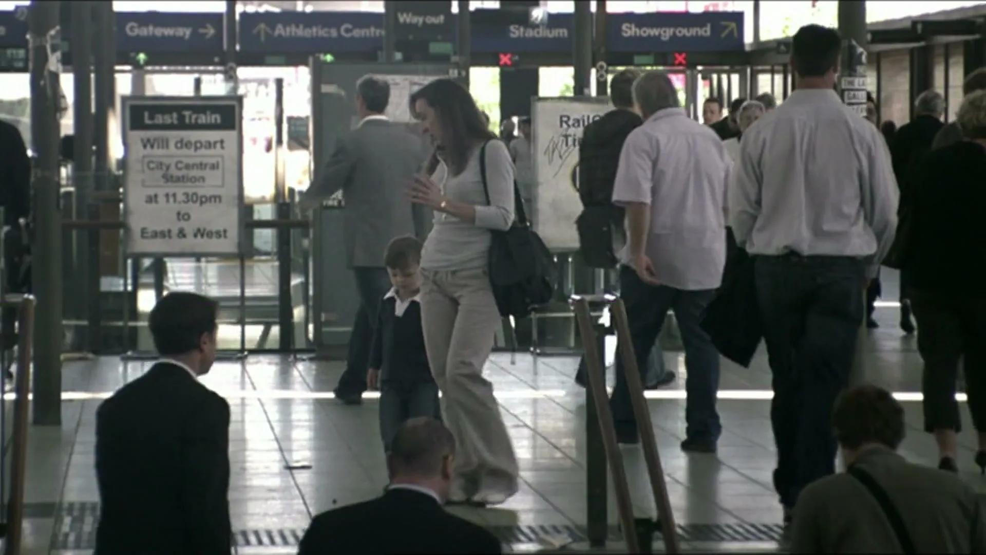 A young boy in a suit holds a woman's hand as they walk through a busy train station, surrounded by other commuters.