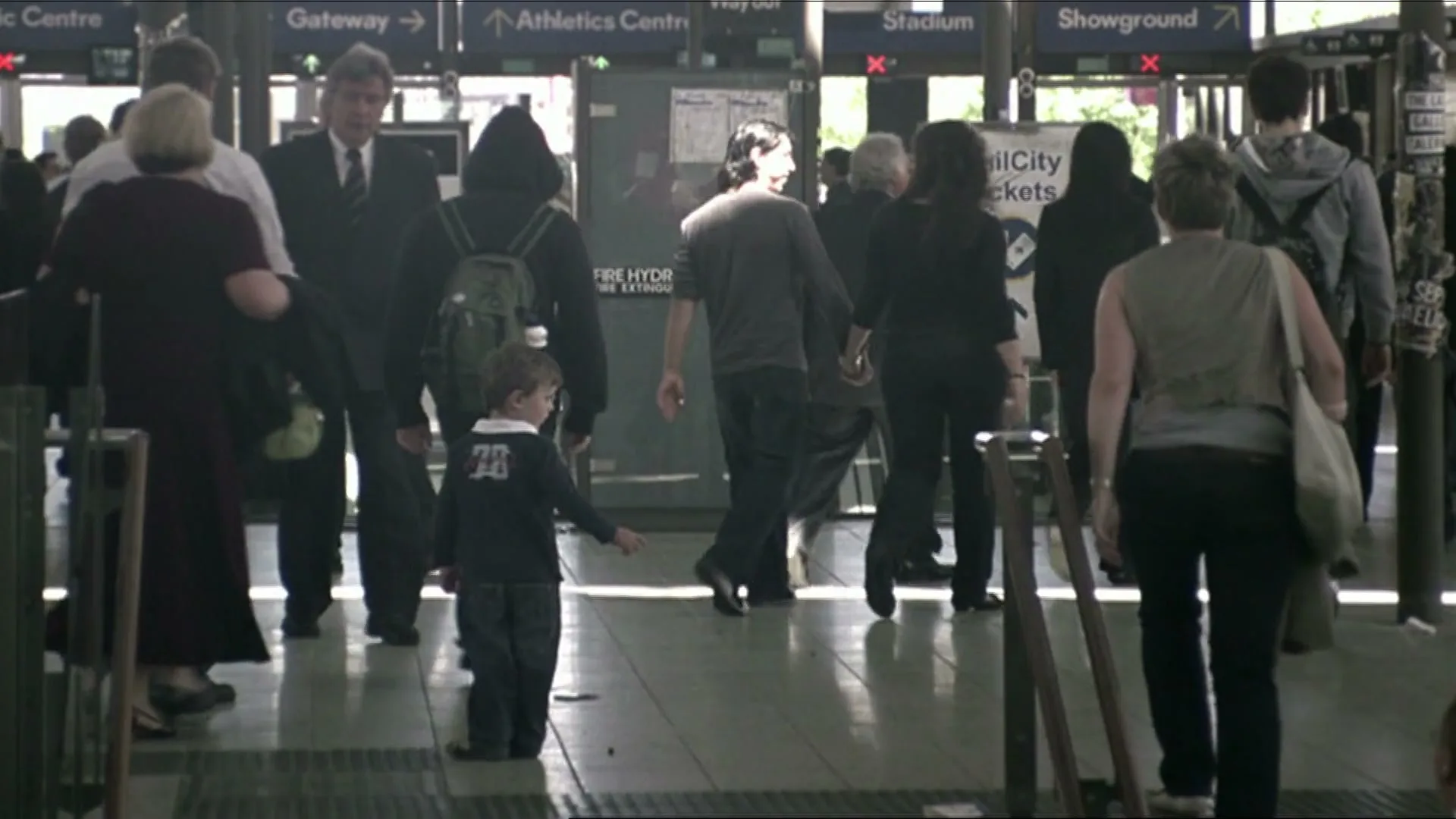 A small boy in a dark shirt stands alone in a busy train station, as people walk past him in various directions.