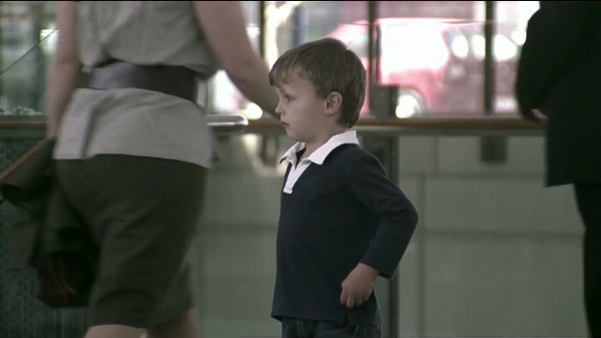 A young boy in a navy blue shirt with a white collar stands still in a busy public space, as adults walk past him.