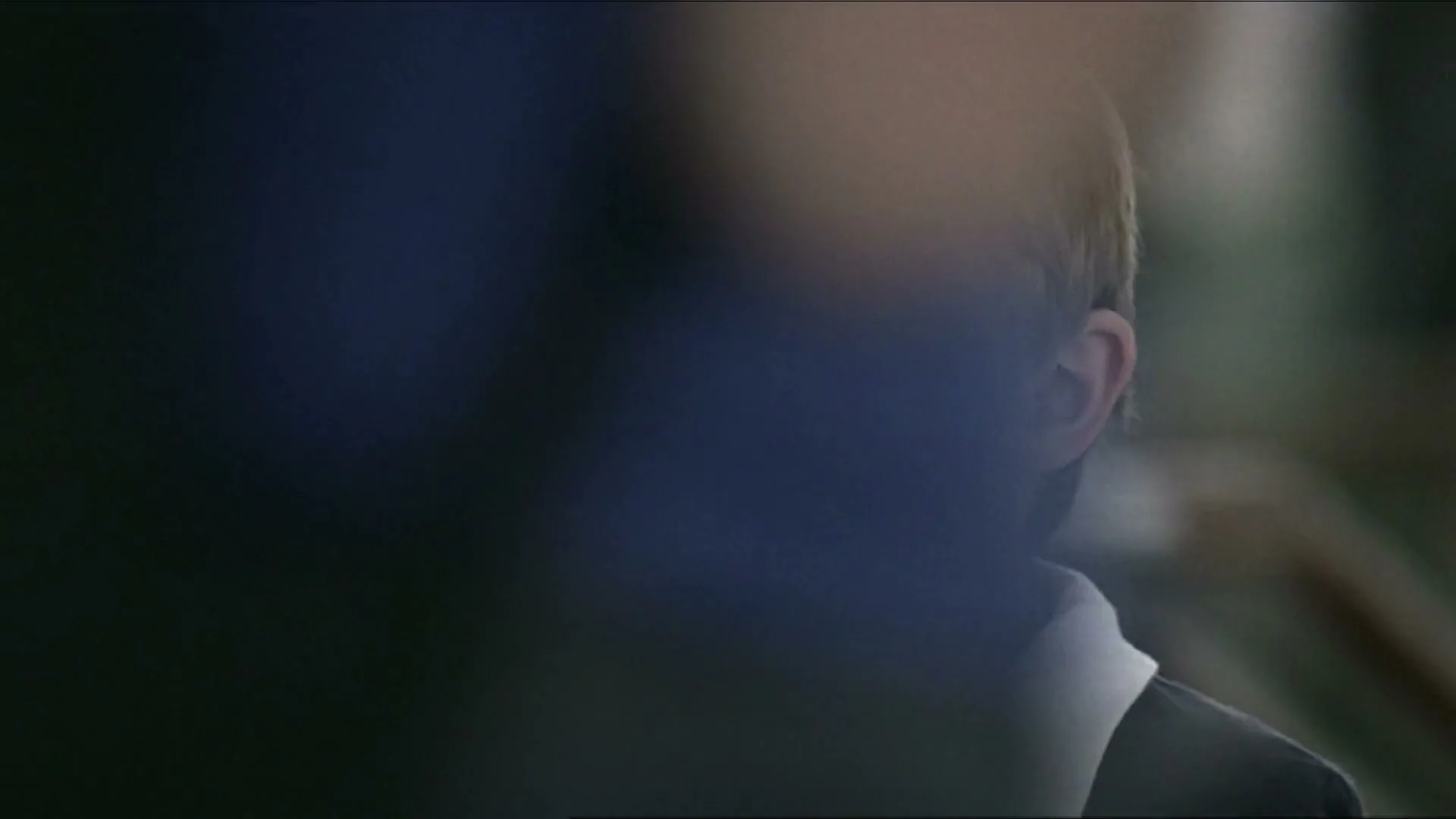 A young boy with brown hair and a white-collared shirt looks around with a sad expression, partially obscured by a dark foreground element.