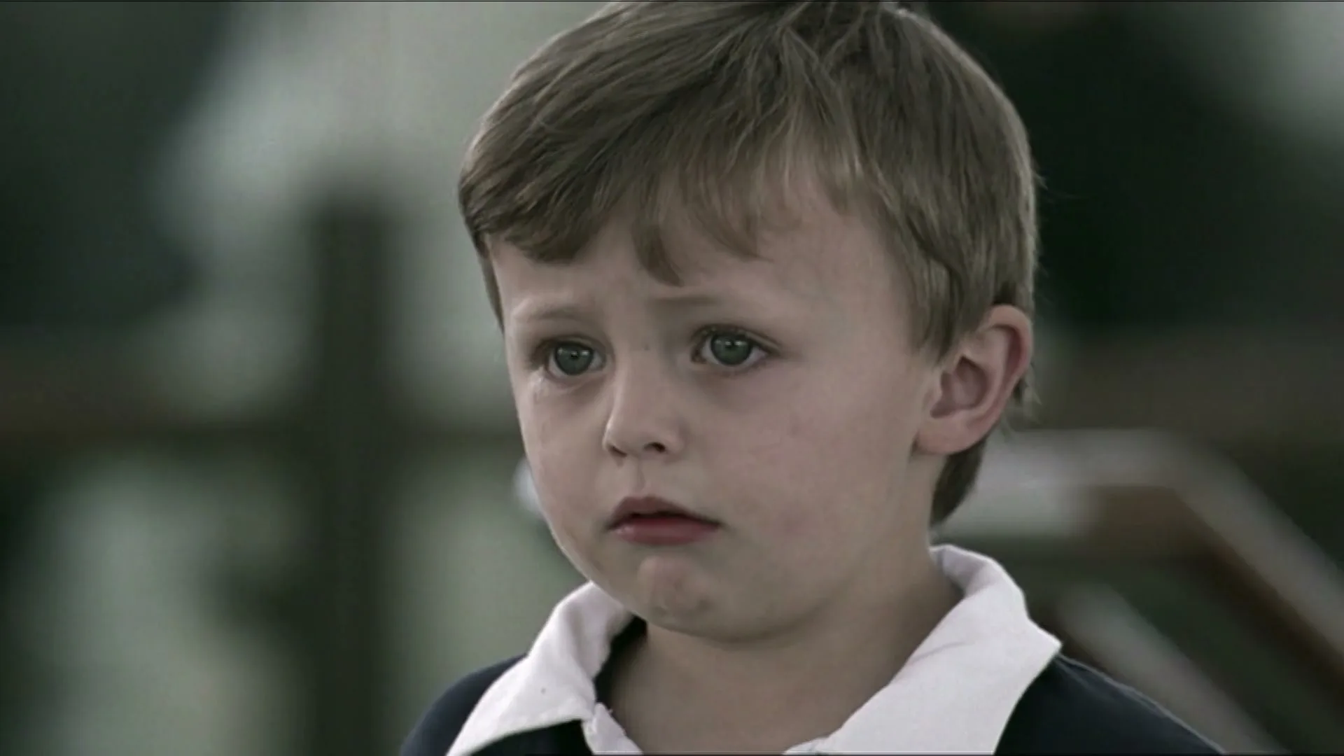 A young boy with brown hair and blue eyes, wearing a navy polo shirt, looks sad and puts his hand to his mouth in an interior setting.
