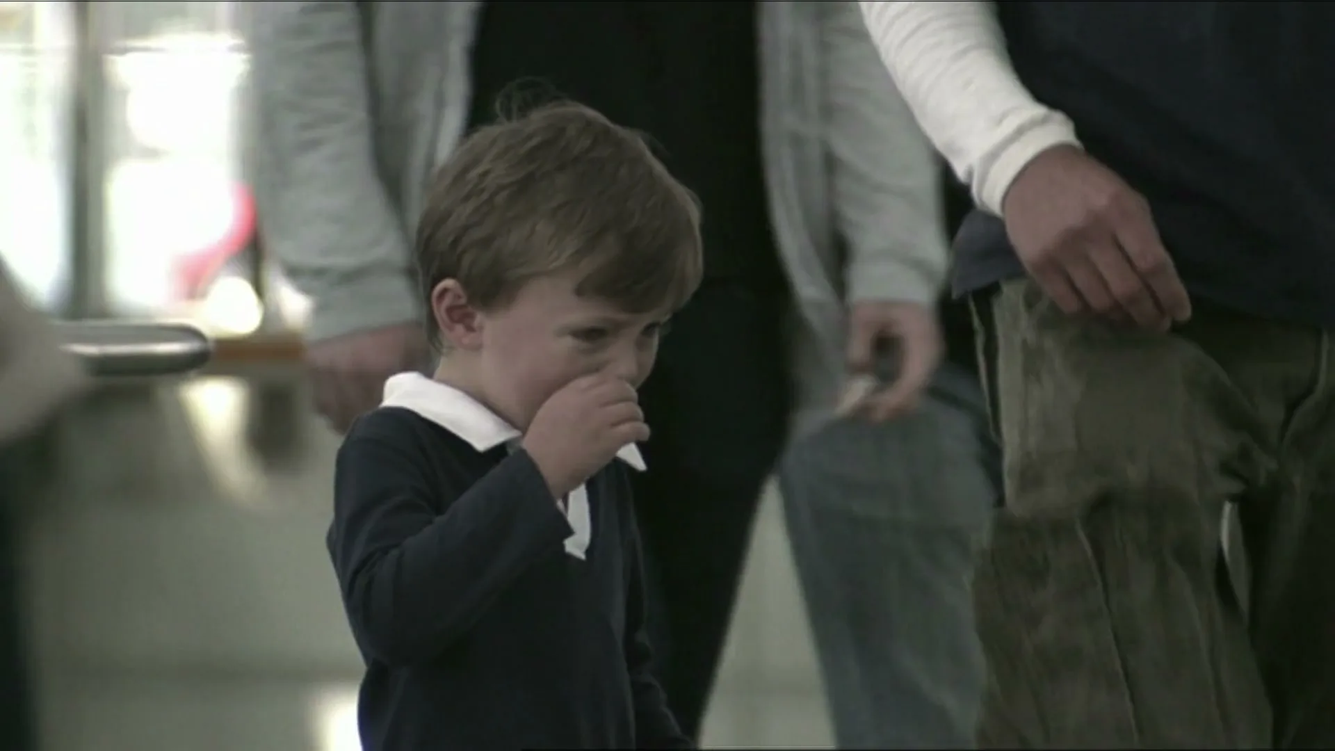 A young boy in a navy polo shirt with a white collar stands amidst a crowd of adults, rubbing his eye as if crying in an interior public space.