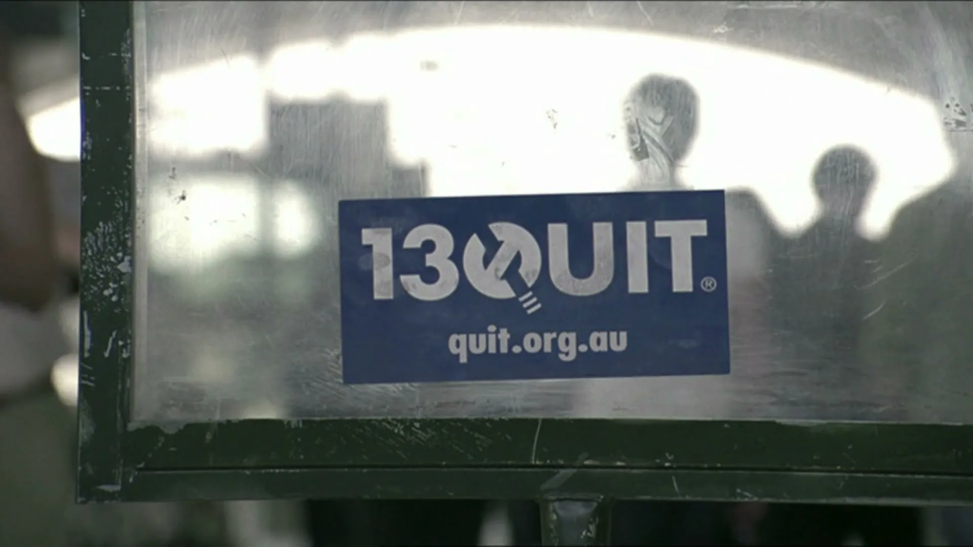 A blue sticker with "13QUIT quit.org.au" is affixed to a dirty glass panel, with blurred silhouettes of people in the background.