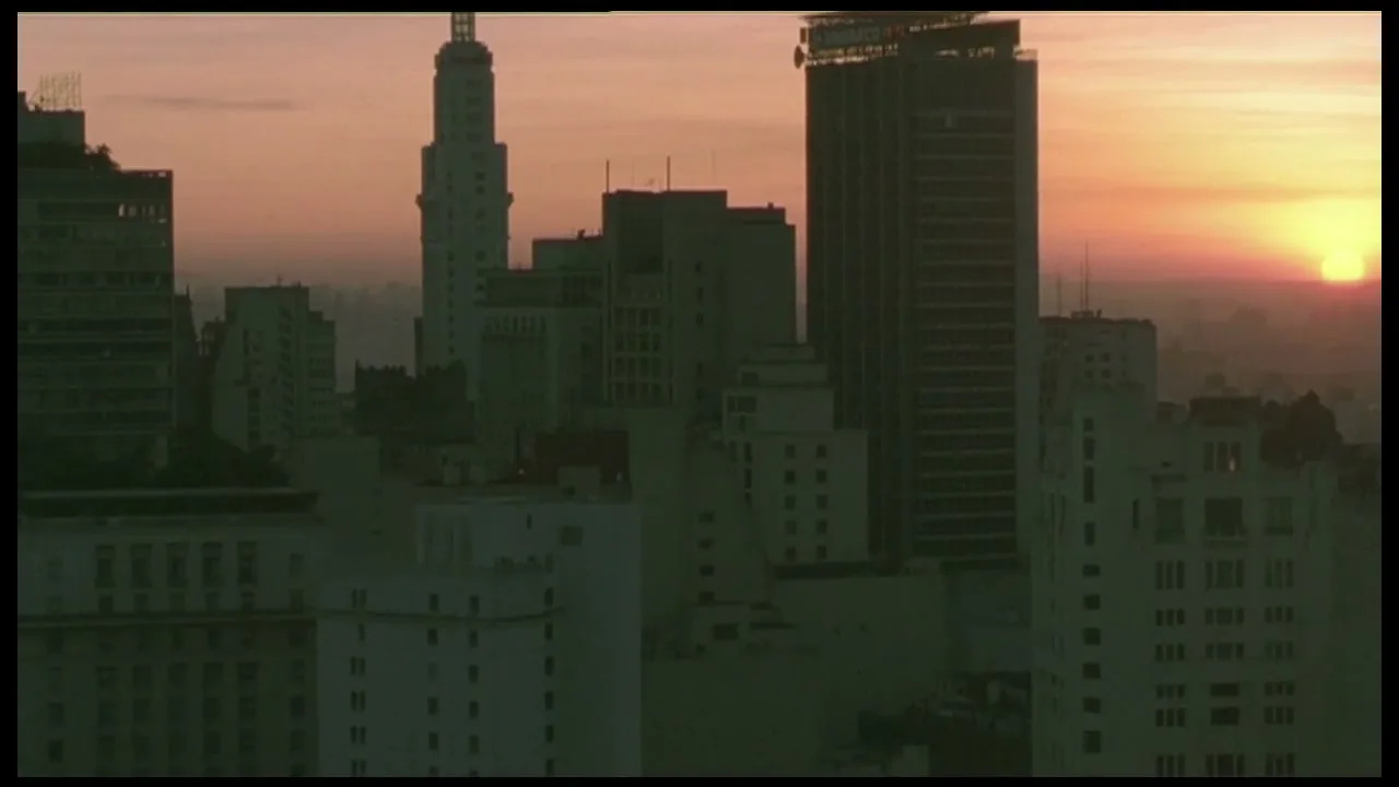 A wide shot of a city skyline at sunrise, with tall buildings silhouetted against the orange and pink sky.