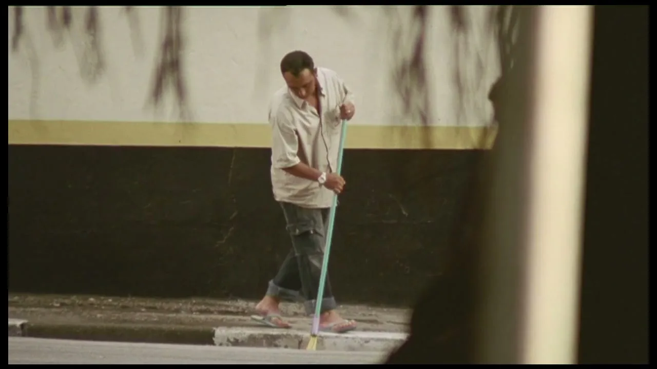 A man in a light shirt and jeans sweeps a sidewalk with a broom, while a boy walks past in the foreground.