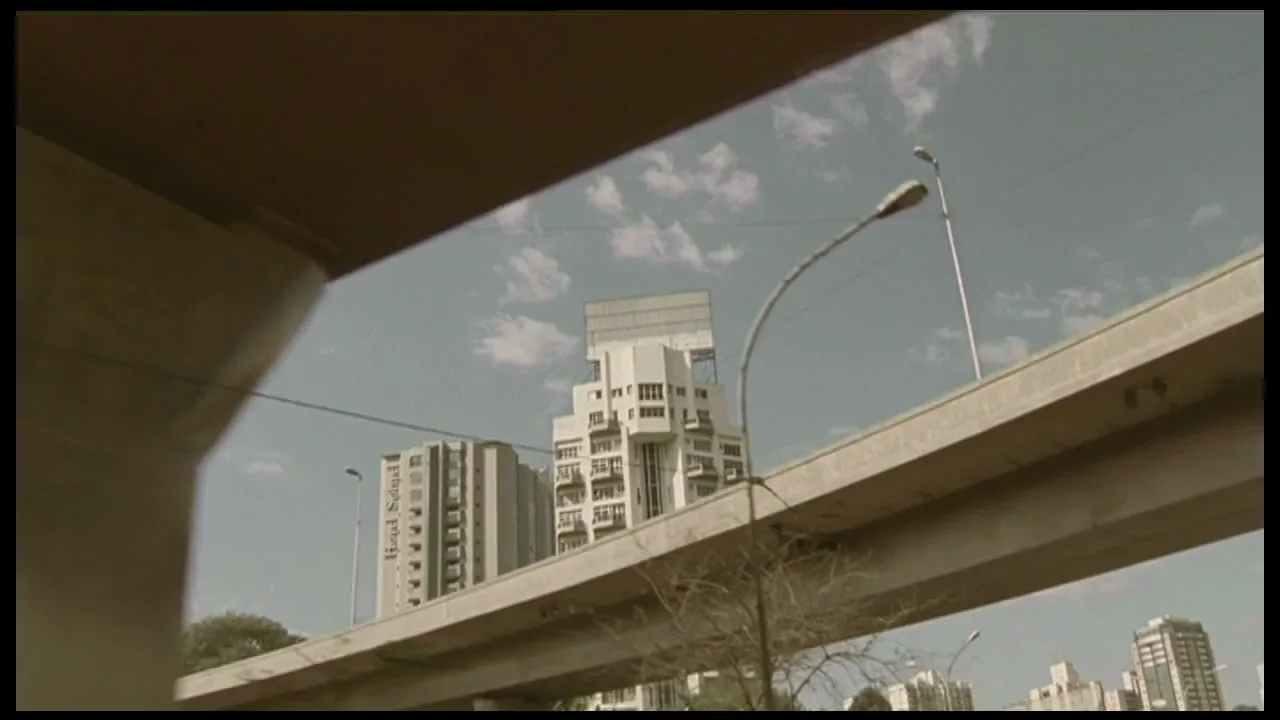 A camera pans right under a highway overpass, revealing city buildings and a blue sky with clouds.