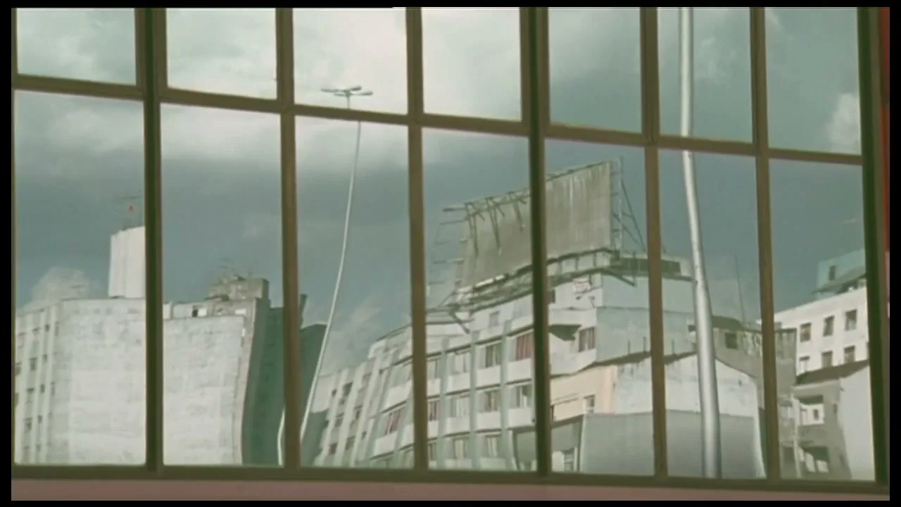 A distorted reflection of buildings and a billboard is seen in a window, with a cloudy sky overhead.