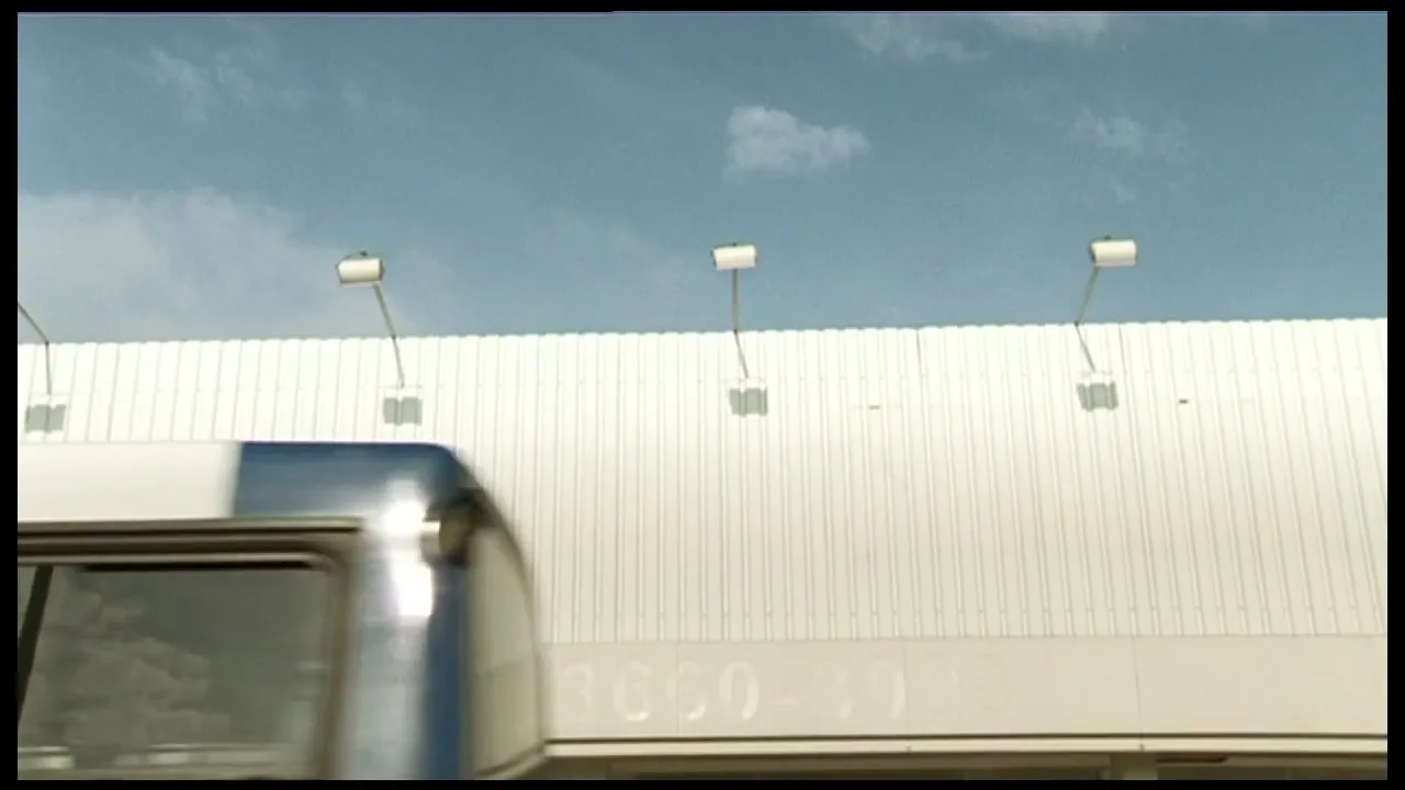 A white billboard with three lights on top is seen against a blue sky as a blue and white bus passes by, reflecting the sky.