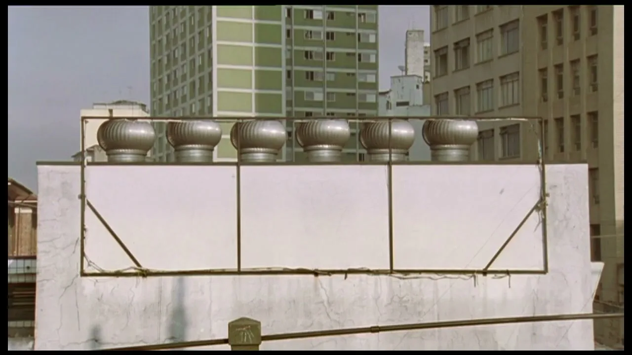 A row of six silver industrial vents sits atop a white building, with other city buildings visible in the background under a bright sky.