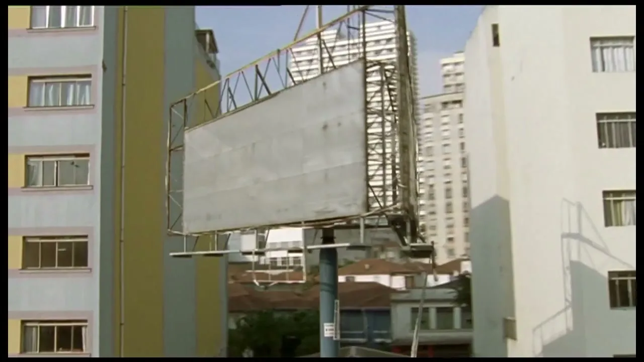 A wide shot of an empty billboard in a city, surrounded by tall buildings and residential houses under a clear sky.