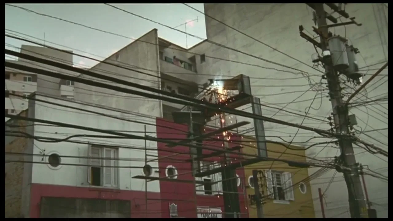 A welder works on a metal structure, creating sparks, in front of colorful buildings and a tangled utility pole in an urban setting.