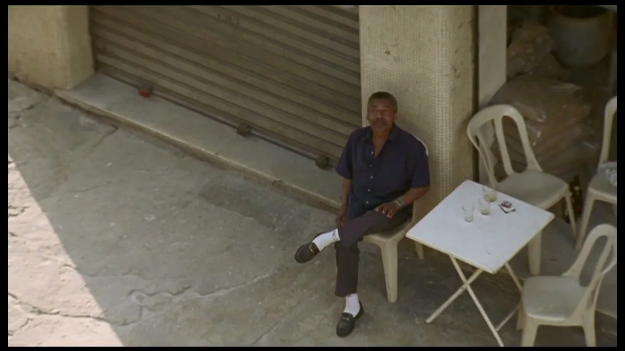 A man in a dark blue shirt and dark pants sits on a white plastic chair at a small white table on a sunny street.