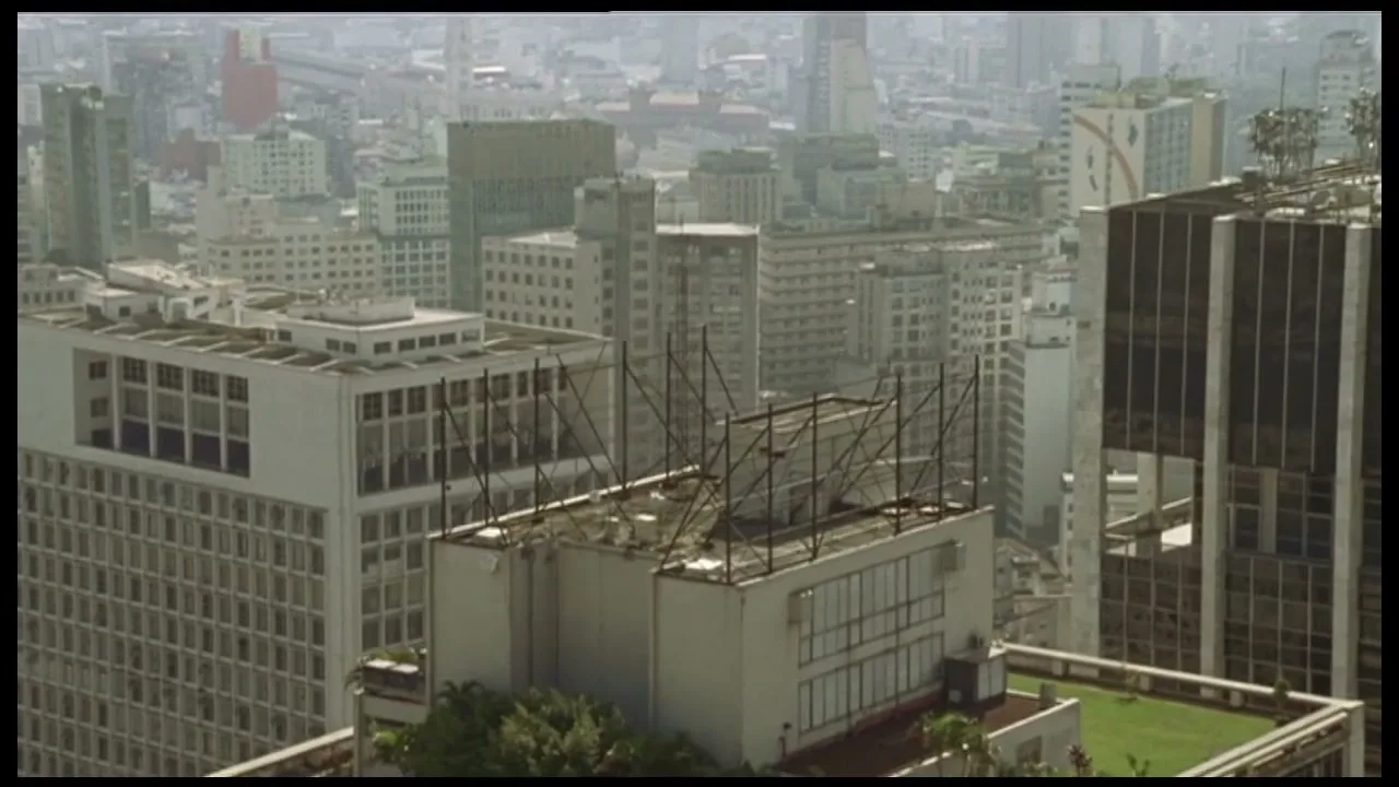 An aerial view of a sprawling city with numerous buildings and empty billboard frames on rooftops under a hazy sky.