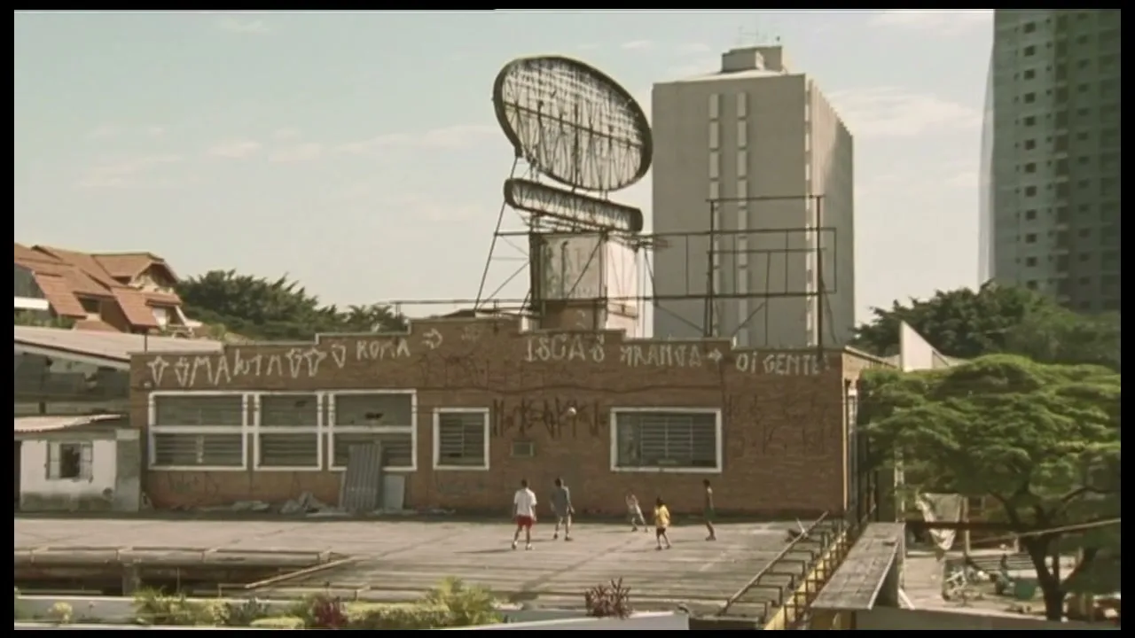 A group of children play soccer on a rooftop in a city, with buildings and trees in the background under a clear sky.