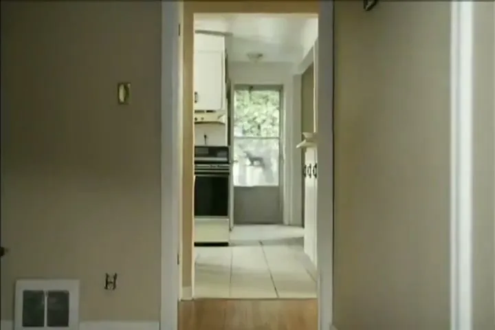A young man in a dark green sweater and brown pants walks through a doorway into a kitchen in a suburban home.