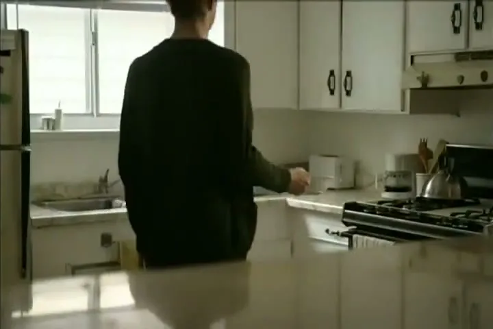 A man in a dark green shirt walks into a kitchen, places a bowl on the counter, and opens a cabinet.