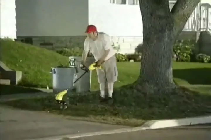 A man in a red hat and white clothes uses a weed trimmer around a tree on a grassy patch next to a sidewalk in front of a house at night.