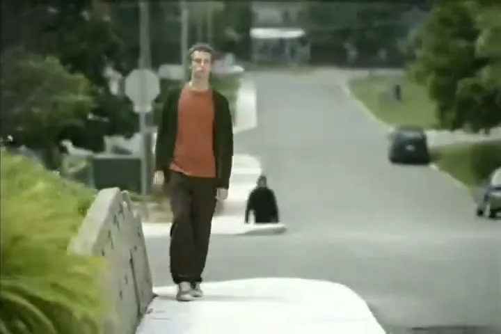 A young man in a red shirt and dark cardigan stands on a sidewalk, looking around as another person walks past him on a suburban street.