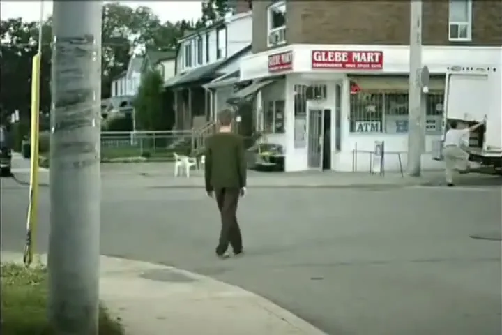 A man in a green suit walks slowly across a street intersection towards a convenience store, with a delivery truck parked nearby.