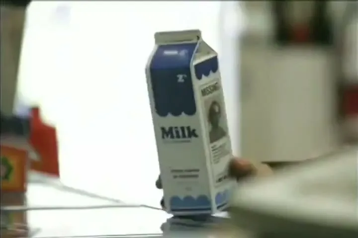 A person's hand places a milk carton with a "missing" person's image on it onto a supermarket checkout conveyor belt.