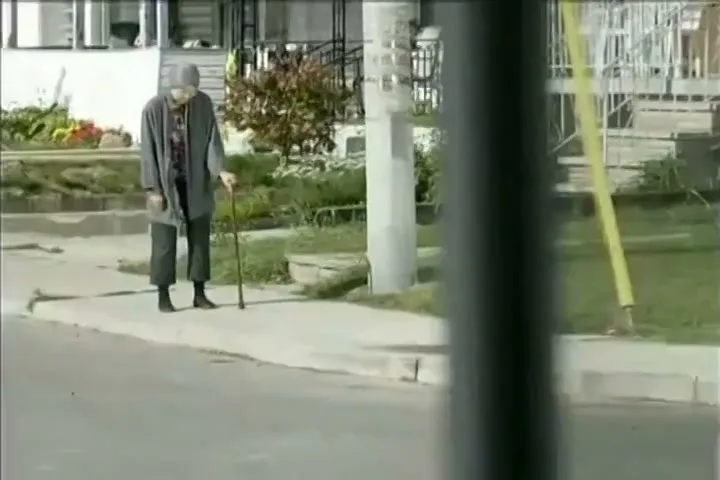 An elderly man with a cane stands on a sidewalk, looking down, as a car drives past him on a sunny day in a suburban neighborhood.