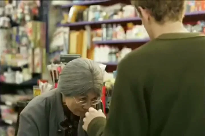 An elderly Asian woman with grey hair and glasses sniffs a carton of milk while a man in a green sweater stands nearby in a convenience store.
