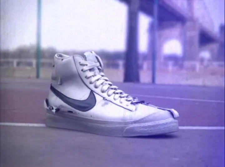 A white Nike Blazer shoe with a black swoosh appears on a basketball court, with a bridge in the blurry background.