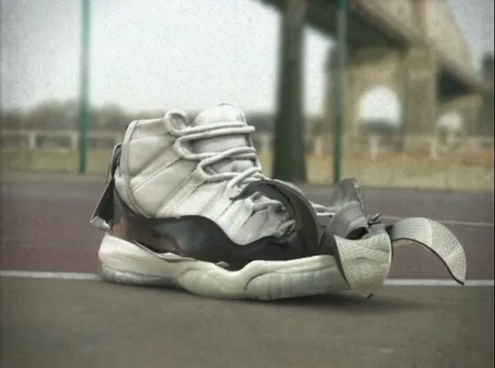 A white and black basketball shoe with a cartoonish toe sits on a basketball court, slowly deforming as if melting.